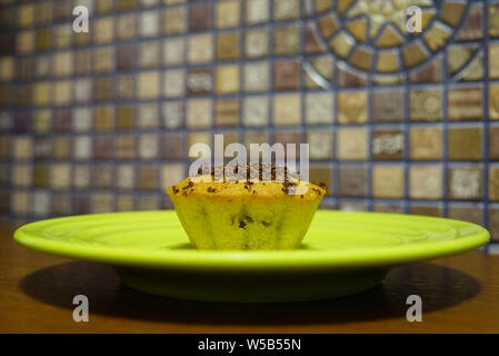 Cupcake mit Chocolate Chips auf einem grünen Untertasse Nahaufnahme. Eine Platte steht auf einem Holz- braun Tabelle. Mosaik Wand im Hintergrund Stockfoto