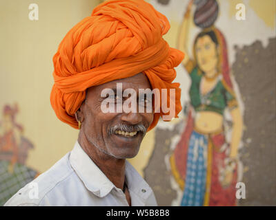 Indische Rajasthani Mann mit einem orangefarbenen Rajasthani Turban (pagari) posiert für die Kamera vor einem Wandbild. Stockfoto