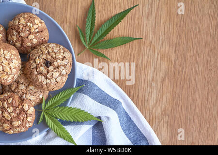 Ansicht von oben ein paar Haferflocken Topf Cookies auf graue Platte auf ein gestreiftes Handtuch auf hellem Holz- Oberfläche und zwei Blätter von Marihuana neben, selektiver Fokus Stockfoto