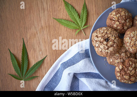 Ansicht von oben ein paar Haferflocken Topf Cookies auf graue Platte auf ein gestreiftes Handtuch auf hellem Holz- Oberfläche und zwei Blätter von Marihuana neben, selektiver Fokus Stockfoto