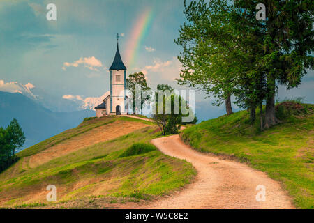 Alpine Landschaft mit Kirche auf dem Hügel. Erstaunlich bunten Regenbogen Landschaft und berühmten Saint Primoz Kirche mit hohen Bergen im Hintergrund, Jamnik Stockfoto