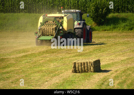 BURTENBACH, BY, Deutschland - Juli 28, 2019: Traktor drücken Sie Heu Quader auf einer Wiese vor einem Kornfeld, drücken Sie die Ballenpresse für Hay Stacks nach havesting Stockfoto