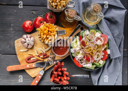 Hausgemachte Pommes Frites und Griechischer Salat. Zwiebeln, Knoblauch, geräuchert Würste, Tomatensauce, Gabel und Löffel auf einem dunklen Holztisch. Ansicht von oben Stockfoto