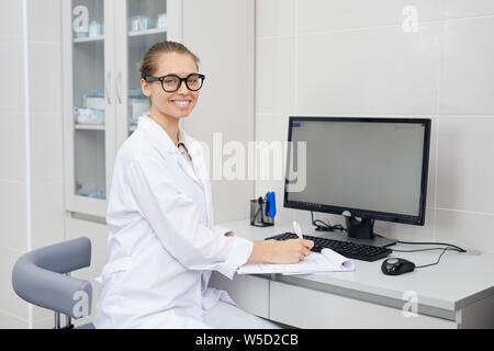 Portrait von hübschen weiblichen Doktor Brille fröhlich in die Kamera lächelt, während die, die sich Notizen am Schreibtisch im Büro, kopieren Raum Stockfoto