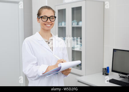 Taille bis Portrait von hübschen Ärztin holding Zwischenablage und fröhlich lächelnd in die Kamera während im Büro Posing, kopieren Raum Stockfoto