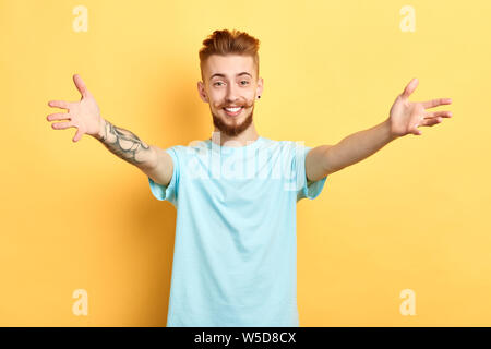 Junge schöne fröhliche Mann, blaues T-Shirt mit offenen Armen für die Hug über isolierte gelben Hintergrund. Herzlich willkommen zu einem Barbershop. Nahaufnahme po Stockfoto