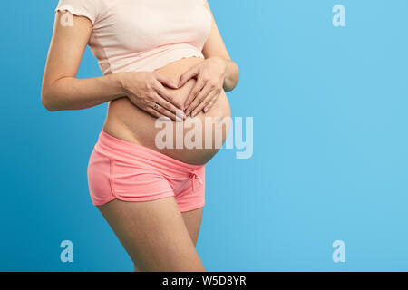 Schwangere Mädchen halten ihre Handflächen im Herzen Form auf ihren Bauch. Nahaufnahme 7/8 Foto. Kopieren Sie Raum. Isolierte blauen Hintergrund. Studio starke warm geschossen. Stockfoto