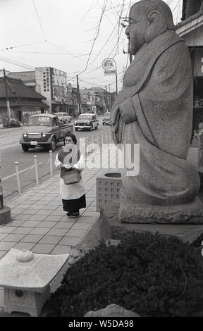 [1950er Jahre Japan - buddhistische Statue und Japanische Hausfrau] - eine Hausfrau, die eine Einkaufstasche und das Tragen einer Schürze und geta ist vor einer Statue von ebisu an Tengenji (天現寺), Tokyo 1957 (Showa 32). Ebisu ist eine der sieben Gottheiten der Fortune (shichi Fukujin). Stockfoto