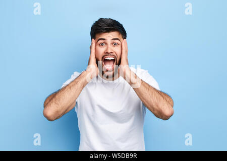 Enttäuscht verwirrt Mann berührt seine Wangen mit Palmen, bringt Verwirrung, lassen Sie sich überraschen. nach oben Porträt schließen. Isolierte blauen Hintergrund, Studio erschossen. neg. Stockfoto