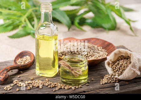 Hanf Öl in Glas und Flasche, Samen in Keramik Teller und Löffel und in Sack auf Holzplatte mit Blättern auf Hintergrund. Stockfoto