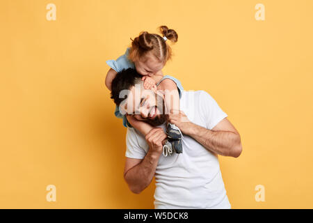 Kleines Mädchen will Papa's Aufmerksamkeit, Schöne Funny Girl kitzeln Ihr älterer Bruder, während er auf seinen Schultern. schließen Foto. Isolierte gelb Bac Stockfoto