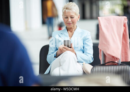 Lächelnde blonde Frau in Blau Jacke sitzen auf Ledersitz im Flughafen und über Wlan auf Minianwendung Stockfoto
