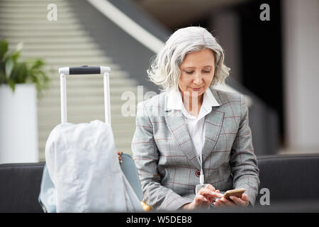 Lächelnde grauhaarige Asiatische Dame in karierten jacke sitzt im Flughafen und Kontrolle Messenger auf dem Telefon Stockfoto
