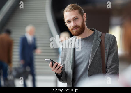 Portrait von positiven Rotbärtigen Kerl in graue Jacke steht in der Lobby und über Gadget Stockfoto