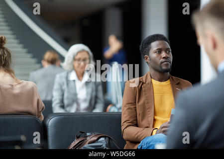 Ernsthafte junge schwarze Mann mit Bart auf Sitz mit Tasche im Flughafen und warten auf Boarding Stockfoto