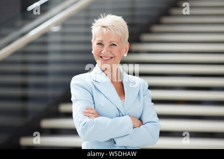 Portrait von fröhlichen attraktive Dame in blau Jacke stand mit verschränkten Armen in der Lobby Stockfoto