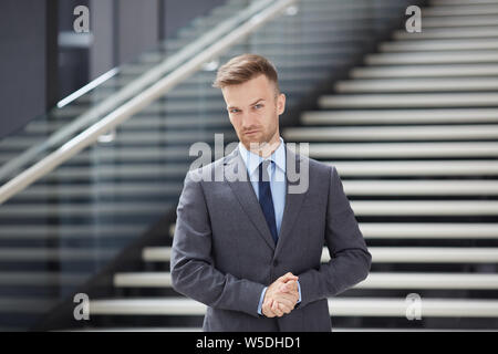 Ernsthafte hübscher junger Geschäftsmann in grauen Anzug gegen Treppe in der Lobby und an der Kamera zuversichtlich Stockfoto