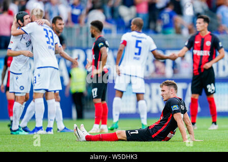 28 Juli 2019, Hessen, Wiesbaden: Fussball: 2. Bundesliga, SV Wehen Wiesbaden - Karlsruher SC, 1. Spieltag, in der BRITA-Arena. Der Wiesbadener Maximilian Dittgen sitzt auf dem Feld nach dem Ende des Spiels enttäuscht, während die Karlsruher Spieler hinter Ihnen ihren Sieg feiern. Foto: Uwe Anspach/dpa - WICHTIGER HINWEIS: In Übereinstimmung mit den Anforderungen der DFL Deutsche Fußball Liga oder der DFB Deutscher Fußball-Bund ist es untersagt, zu verwenden oder verwendet Fotos im Stadion und/oder das Spiel in Form von Bildern und/oder Videos - wie Foto Sequenzen getroffen haben. Stockfoto