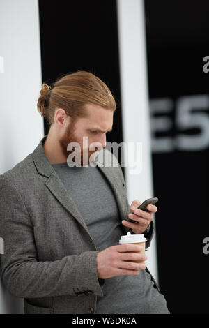 Red konzentriert - bärtigen Mann in Jacke lehnte sich an die Wand und Lesen von SMS auf dem Smartphone beim Trinken von Kaffee in der Lobby Stockfoto