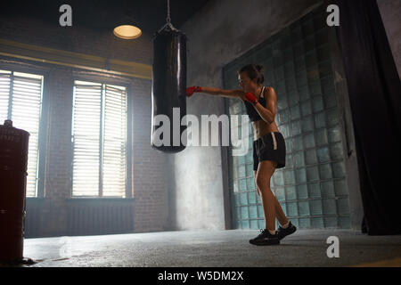 Dramatische Seitenansicht Portrait von harten Kämpferin schlagen Boxsack boxing Praxis, kopieren Raum Stockfoto