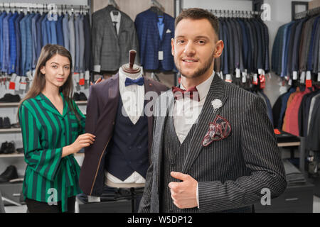 Junger Mann mit Bart im eleganten Anzug in Fashion Boutique posiert, an der Kamera schaut. Zuversichtlich Geschäftsmann Jacke tragen, weißes Hemd, Weste und roter Fliege. Shop Berater in der Boutique. Stockfoto