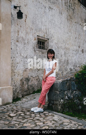 Junge lächelnde Frau Touristen sitzen auf einer Burg von Bled Wand im Innenhof. Tragen weiße Hemd, weiße Turnschuhe und Koralle Hose. Travel Concept Stockfoto