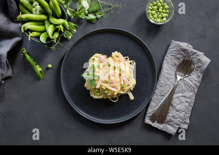 Pasta Tagliatelle mit Sahne, Erbsen und Schinken auf schwarzem Hintergrund. Köstliche mediterrane Mittagessen. Ansicht von oben. Stockfoto