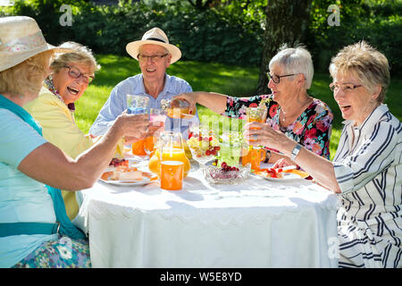 Gerne ältere Menschen um den Tisch Picknick sitzen. Gruppe von Senioren Obst Kuchen essen und trinken Orangensaft, eine Frau ist Saft serviert. Stockfoto