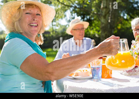 Senioren mit Picknick im Freien bei Sonnenschein, blonde schöne Frau mit Hut ist mit Orangensaft zu alten Freunden. Stockfoto