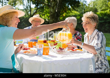 Happy Gruppe von älteren Freunden picknicken im Park, Kuchen essen und Obst, Kaffee trinken, blonde schöne Frau mit Hut ist mit Orangensaft. Stockfoto