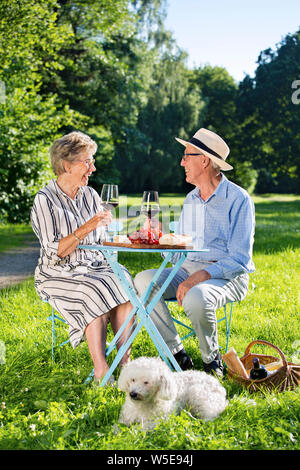 Gerne älteres Paar einander in die Augen schauen. Am Tisch sitzt, niedlichen weißen Hund zu ihren Füßen, Picknicken im Park, Wein trinken, essen Käse wi Stockfoto
