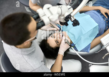 Männliche Zahnarzt in eine graue Uniform mit Schutzmaske und weiß Latex Handschuhe sieht in einer zahnmedizinischen Mikroskop während seine Patientin in einem blauen Bib behandelt Stockfoto