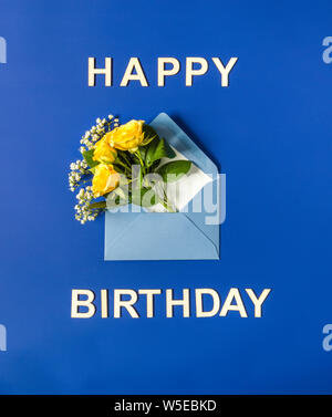 Gelbe Rosen und weißen Gypsophila in blauen Umschlag close-up auf blauem Hintergrund. Text Happy Birthday, Holz- briefe. Ansicht von oben, flach. Vorlage für Stockfoto