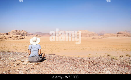 Einsame Frau mit Hut sitzt auf dem Boden mit Blick auf die Wüste des Wadi Rum in der Kontemplation. Stockfoto