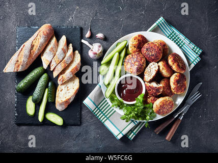 Ansicht von oben der Polnischen Fleisch Kuchen serviert mit Gurkenscheiben und Barbeque Sauce auf eine weisse Platte auf einer konkreten Tabelle mit geschnittenem Baguette, Ho Stockfoto