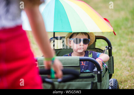 Ein Kind mit Sonnenbrille und Sonnenhut sitzt in einem Trolley als seine Familie kommt auf einem Festival, Großbritannien Stockfoto