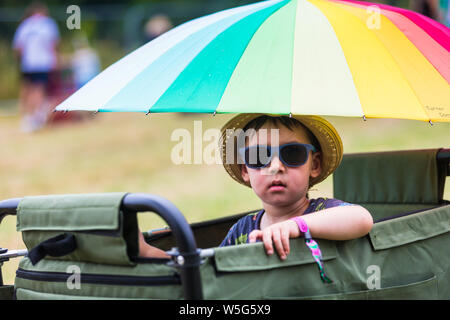 Ein Kind mit Sonnenbrille und Sonnenhut sitzt in einem Trolley als seine Familie kommt auf einem Festival, Großbritannien Stockfoto