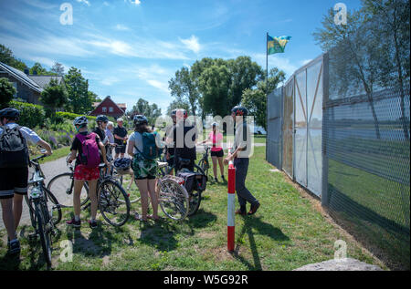 18. Juni 2019, Mecklenburg-Vorpommern, Rüterberg: Andreas Wagner, Mitarbeiter der Politische Memoriale e.V. und Leiter des Grenzhuus, erklärt die 30 Radfahrer auf Ihrer einwöchigen Tour entlang der ehemaligen innerdeutschen Grenze ein erhaltenes Stück Grenzzaun im Dorf Republik Rüterberg. Aufgrund seiner exponierten Lage in der DDR direkt an der westlichen Grenze, Rüterberg wurde komplett von einem drei Meter hohen Zaun umgeben. Einen Tag vor dem Fall der Berliner Mauer am 8. November 1989, die Bewohner in einer Sitzung gefordert, die "Öffnung der Grenzen der DDR" und erklärten ihre Stockfoto