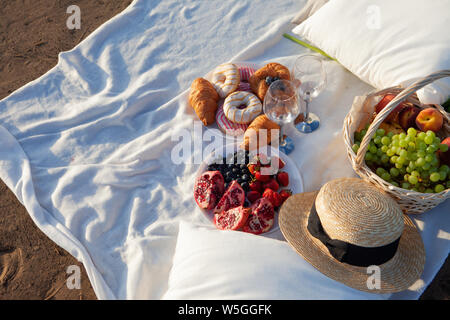 Picknick in den Strahlen der untergehenden Sonne auf den Sand. Auf der Plaid liegt ein Korb mit Obst, Gläser und süßem Gebäck. In der Nähe befindet sich ein Strohhut. Stockfoto