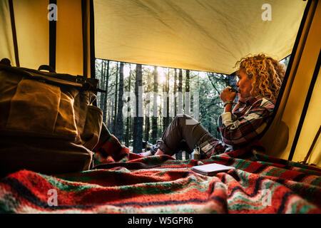 Unabhängige Schöne lockige blonde Haare Frau sitzen vor einem Zelt im Freien wildes Campen im Wald genießen eine Tasse Kaffee und Denken - Outdoor Stockfoto