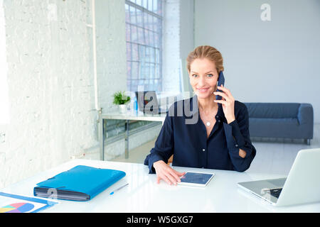 Portraitfotos der mittleren Alters Geschäftsfrau im Gespräch über Ihr Mobiltelefon beim Sitzen im Büro Schreibtisch und funktioniert. Stockfoto