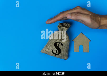 Home Versicherungen. Haus zu schützen. Ein Mann Hand geschützt ein kleines Haus und Geld Tasche über ein Dach auf blauem Hintergrund. Stockfoto