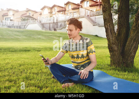 Junge stilvolle hübscher Kerl sitzt auf Gras und Telefonieren im Park ausruhen. Pause Stockfoto