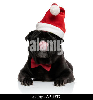 Jolly schwarz pug Blick in die Kamera und leckte sich die Nase beim Tragen eines roten bowtie und ein Weihnachtsmann hat, liegen auf weißem Studiohintergrund Stockfoto