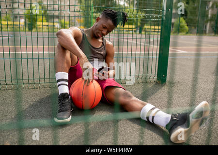 Junge afrikanische Basketballspieler mit Smartphone Entspannen durch Zaun Spielplatz Stockfoto