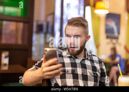 Junge hipster Mann trinken Kakao aus einem Strohhalm schockiert und Nachrichten in sozialen Netzwerken, während an einem Tisch in einem Cafe sitzen. Das Konzept der Stockfoto