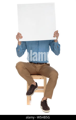 Der moderne Mensch sitzt in Blue Jeans Hemd bedeckt sein Gesicht hinter einem leeren Plakat auf weißem Hintergrund Stockfoto