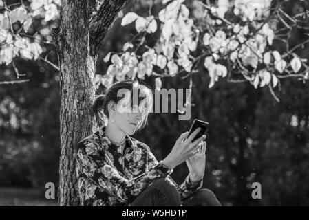 Graustufen Bild einer jungen Frau unter einem Herbst Baum auf Ihrem Smartphone suchen sitzen. Stockfoto