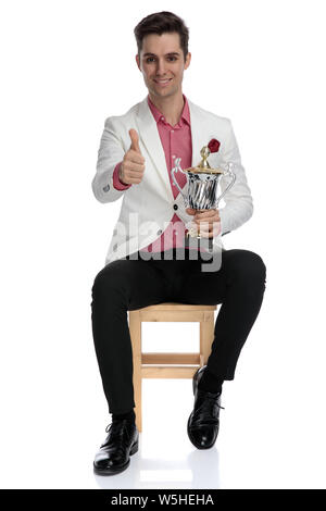 Sitzen junge Smart Casual Mann mit Pokal und macht das ok Zeichen auf weißem Hintergrund Stockfoto