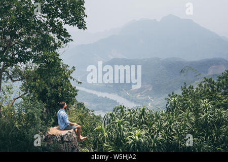 Jungen kaukasischen Mann sitzt oben auf dem Berg mit Blick auf das Flusstal in Nong Khiaw Dorf, Laos Stockfoto